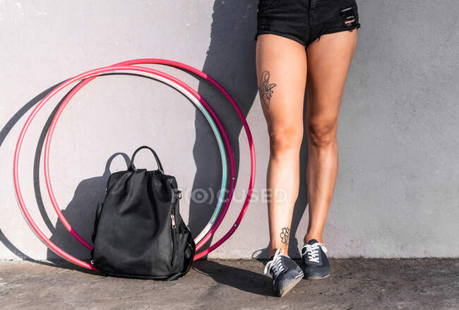 Crop unrecognizable woman in shorts and sneakers standing on pavement with rucksack and hula hoops in sunlight on gray background — Stock Photo