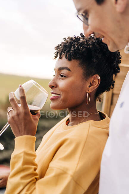 Vista laterale dell'uomo che abbraccia la donna nera in piedi con un bicchiere di vino sul balcone mentre si gode il fine settimana e distoglie lo sguardo — Foto stock