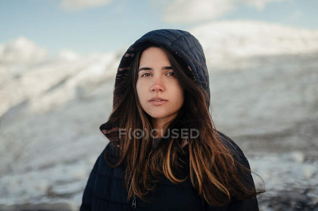 Молода ніжна жінка-мандрівник в капюшоні дивиться на камеру проти засніжених гір взимку в сонячний день — стокове фото