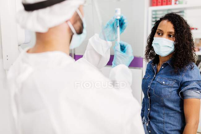 Médecin en gants de latex et bouclier facial remplissant la seringue du flacon avec un vaccin se préparant à vacciner une patiente afro-américaine méconnaissable en clinique pendant l'épidémie de coronavirus — Photo de stock