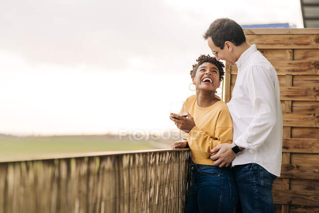 Vista laterale dell'uomo che abbraccia la donna nera in piedi con un bicchiere di vino sul balcone mentre si gode il fine settimana e si guarda — Foto stock