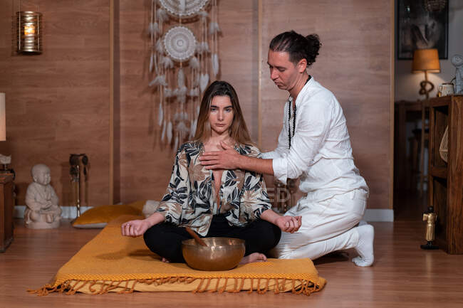 Profesora zen en ropa blanca tocando pecho de mujer con piernas cruzadas durante la práctica de yoga cerca del bowl gong - foto de stock