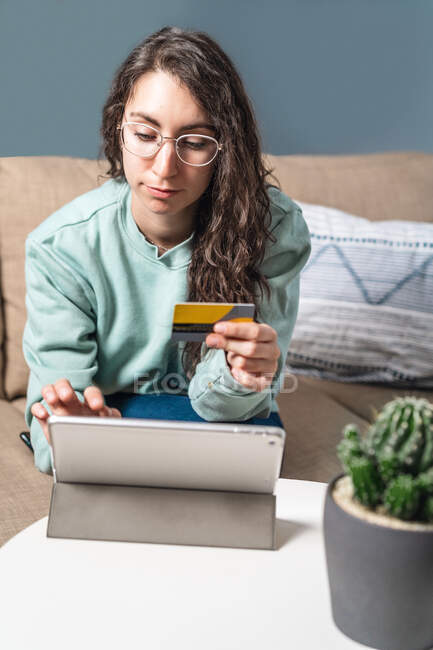 Joven mujer feliz usando su tarjeta de crédito amarilla para comprar en línea con la tableta, sentado en el sofá en casa - foto de stock