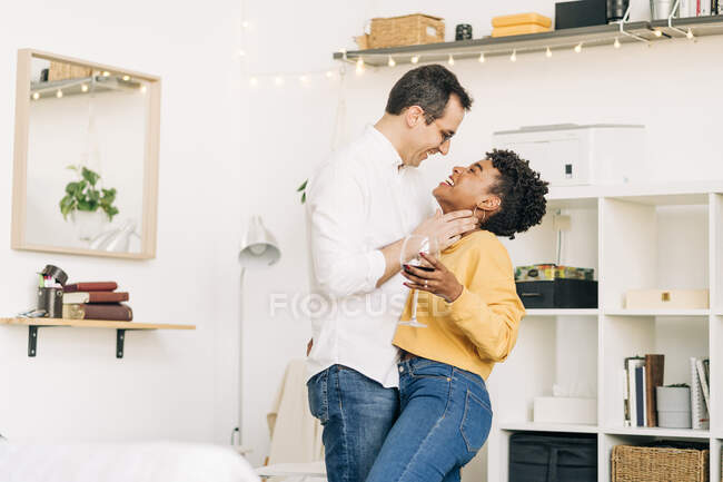 Vista lateral de la sonriente pareja multiétnica enamorada de pie con una copa de vino y abrazada mientras pasaban un fin de semana romántico en casa y se miraban - foto de stock