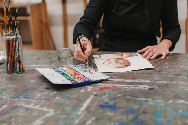 Crop artista femenina en la pintura de delantal con acuarelas en papel mientras está sentado en la mesa en el taller creativo - foto de stock