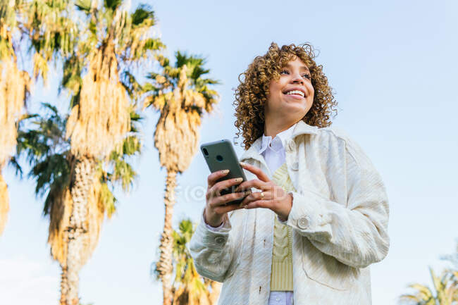 Low angle of cheerful African American female browsing mobile phone while standing in exotic park with palm trees at sunset and looking away — Stock Photo