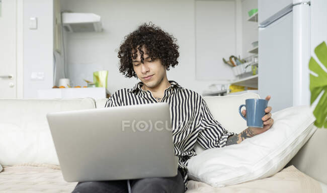 Молодий чоловік-хіпстер з кучерявим волоссям, що переглядає Інтернет на нетбуці, відпочиваючи на дивані в кімнаті — стокове фото