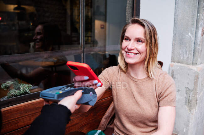 Smiling adult female cafeteria customer paying for hot drink to go with cellphone app through payment terminal against faceless employee — Stock Photo