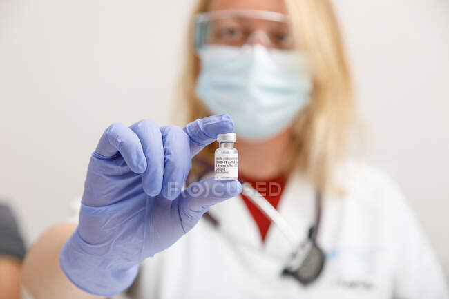 Médico femenino con mascarilla protectora y guantes de látex con vial de vacuna contra el coronavirus que se muestra a la cámara mientras está de pie en la habitación del hospital - foto de stock