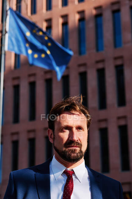 Unshaven executivo masculino de terno com gravata olhando para longe contra a construção de vários andares e bandeira da União Europeia na cidade — Fotografia de Stock
