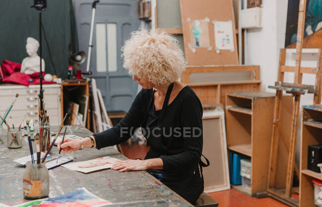 Künstlerin in Schürzenmalerei mit Aquarellen auf Papier, während sie am Tisch in der Kreativwerkstatt sitzt — Stockfoto