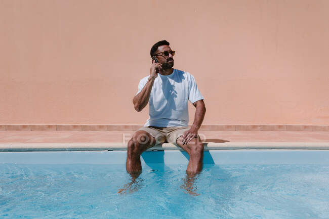Freelancer masculino serio sentado junto a la piscina con las piernas en el agua y hablando por teléfono móvil durante el trabajo remoto en verano - foto de stock