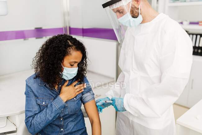 Vista lateral del especialista médico masculino en uniforme protector, guantes de látex y protector facial vacunando a la paciente afroamericana en la clínica durante el brote de coronavirus - foto de stock