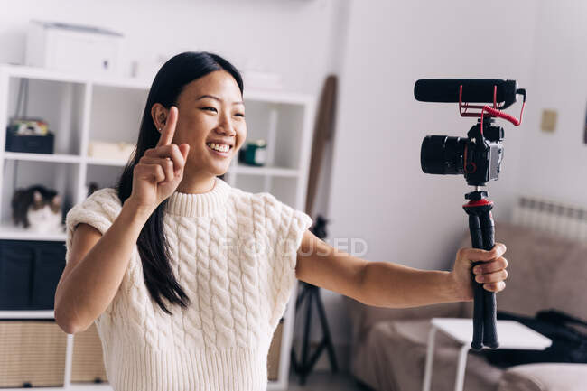 Sorridente vlogger etnico femminile che registra video sulla macchina fotografica mentre fa gesti di mano in piedi in salotto — Foto stock