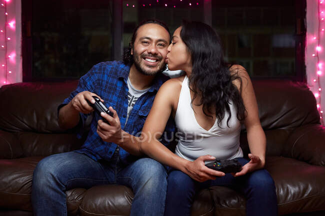 Aufgeregtes ethnisches Paar in Freizeitkleidung mit Joy Pads beim gemeinsamen Spielen von Videospielen, während es zu Hause auf der Ledercouch sitzt — Stockfoto