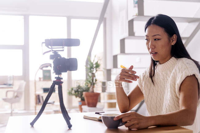 Giovane vlogger etnico femminile con taccuino seduto a tavola con macchina fotografica su treppiede in cucina — Foto stock