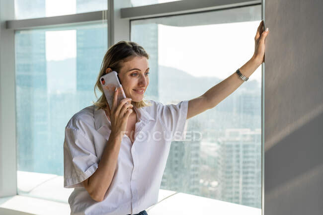 Молода щаслива бізнес-леді, що стоїть в офісі з великими вікнами, має телефонний дзвінок на мобільний телефон — стокове фото