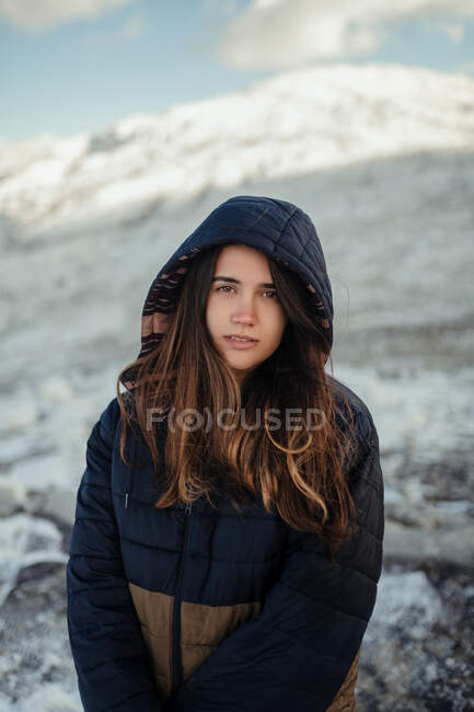 Молодая нежная женщина-путешественница в капюшоне смотрит в камеру на снежные горы зимой в солнечный день — стоковое фото