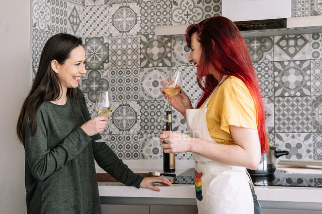 Вид сбоку на молодых улыбающихся гомосексуальных женщин в очках и бутылке белого вина, говорящих в доме, глядя друг на друга — стоковое фото