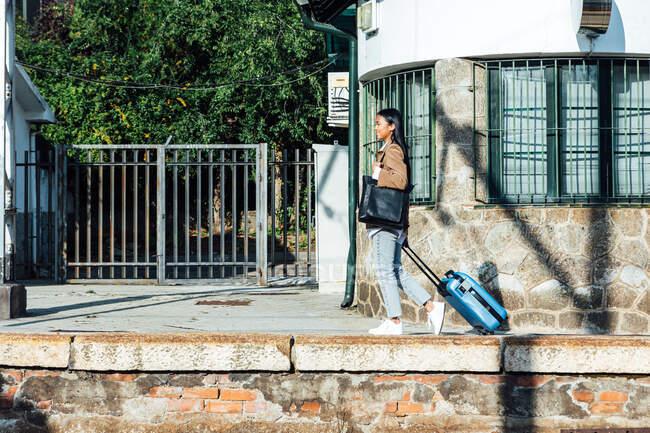 Vue latérale de voyageuse heureuse avec valise marchant le long de la plate-forme à la gare le jour ensoleillé — Photo de stock