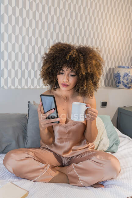 Молодая этническая женщина в спальной одежде с кружкой горячего напитка смс сообщения на мобильный телефон, сидя со скрещенными ногами в спальне — стоковое фото