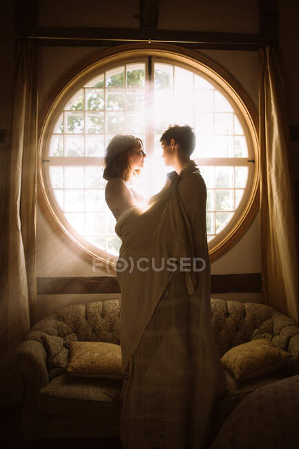 Анонимный мужчина обнимает девушку с текстилем на диване, глядя друг на друга на круглые формы окна в солнечном свете — стоковое фото