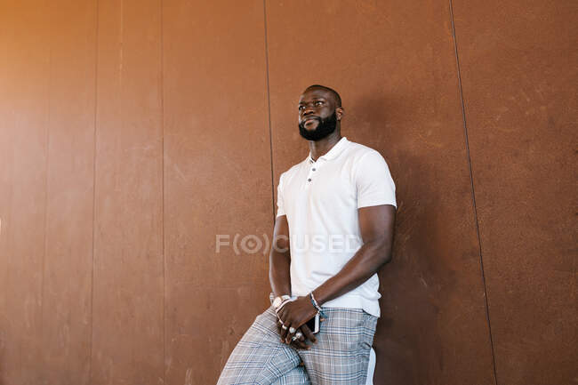 Varón afroamericano barbudo con camiseta blanca y anillos en los dedos de pie con teléfono celular y mirando hacia adelante - foto de stock