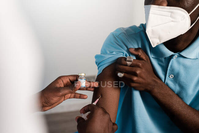 Schwarzer Arzt in Schutzuniform und Latexhandschuhen impft während des Coronavirus-Ausbruchs bis zur Unkenntlichkeit männlichen Afroamerikaners in Klinik — Stockfoto