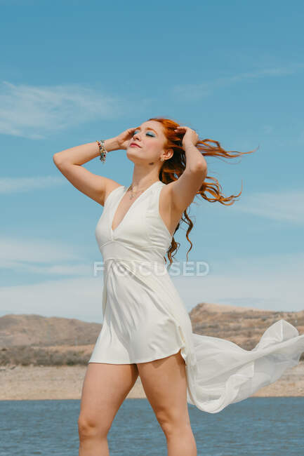 Молода ніжна жінка з макіяжем і закритими очима в білій літаючій сукні торкається червоного волосся проти річки під блакитним хмарним небом — стокове фото