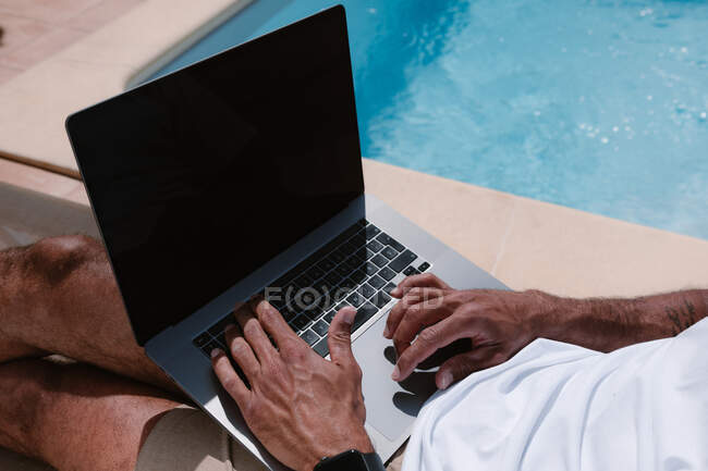 Crop unrecognizable male freelancer lying on lounger at poolside and surfing Internet on laptop during telework in summer on sunny day — Stock Photo