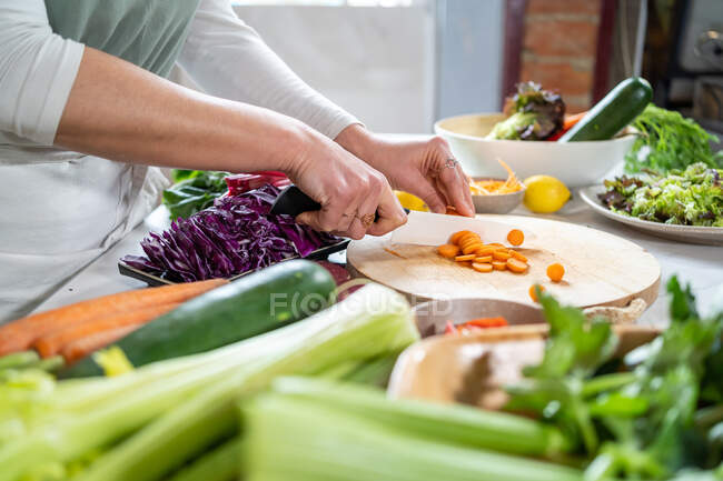 Растениеводство неузнаваемая женщина режет сырую морковь ножом во время приготовления вегетарианской пищи в доме — стоковое фото