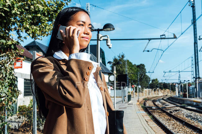 De baixo vista lateral de conteúdo étnico viajante feminino em pé na plataforma na estação ferroviária e falando no smartphone — Fotografia de Stock