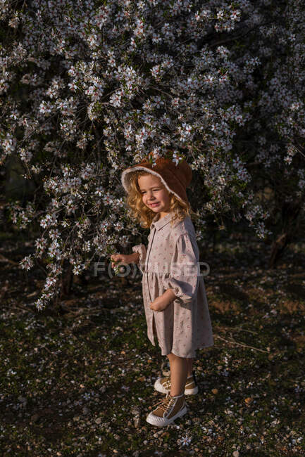 Criança sorrindo adorável no vestido que está perto da árvore florescente com flores no parque da mola e olhando afastado — Fotografia de Stock