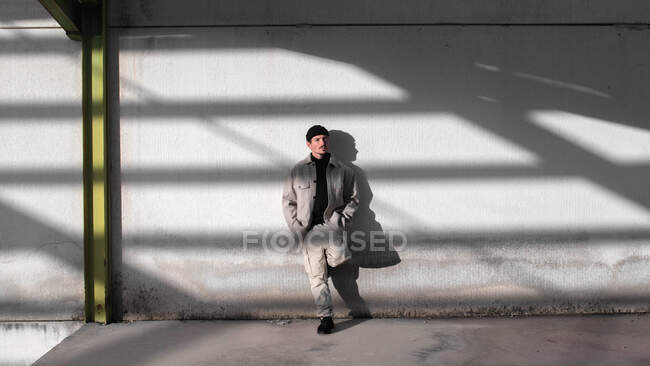 Elegante maschio appoggiato su un muro grigio squallido nella zona urbana della città illuminata dalla luce del sole e distogliendo lo sguardo — Foto stock