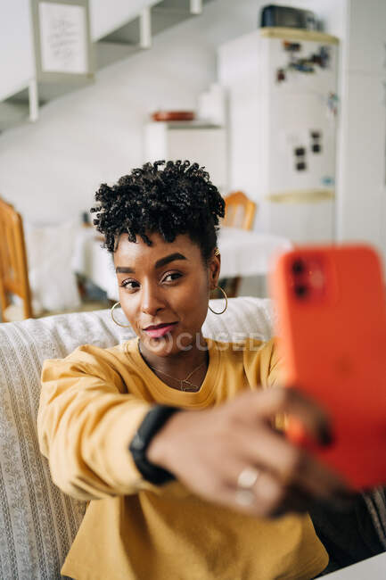 Affascinante donna afroamericana con i capelli ricci che si autoritratto sullo smartphone mentre si siede sul divano di casa — Foto stock