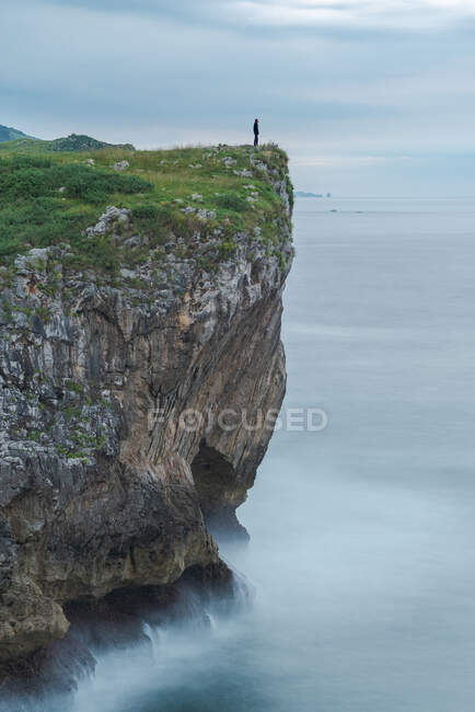 Silueta de persona parada al borde del acantilado rocoso cerca del mar en la costa de Ribadesella en día nublado en Asturias - foto de stock