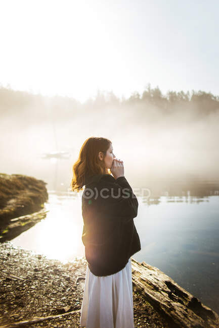 Mujer de pie vestida con un vestido blanco y una chaqueta sobre ella en una roca mirando a un lago en un día de niebla - foto de stock