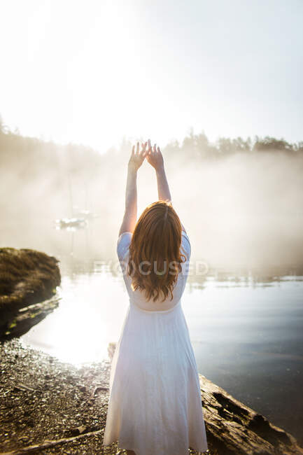 Mujer vista trasera vestida con un vestido blanco en una roca mirando a un lago en un día de niebla - foto de stock