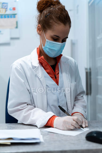 Серйозна жінка-лікар в масці і форма сидить за столом в медичній кімнаті і пише рецепт на папері — стокове фото