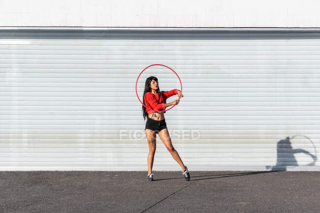 Jeune femme tatouée en vêtements de sport tournoyant hula hoop tout en dansant contre des murs de briques avec des ombres — Photo de stock