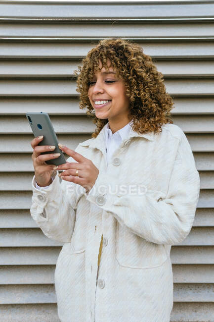 Mulher afro-americana otimista com penteado afro navegando no smartphone enquanto está contra a parede de metal na área urbana da cidade — Fotografia de Stock