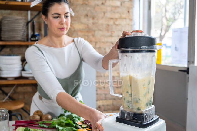 Кукурудзяне жіноче змішування листя з вегетаріанським молоком в кухонному приладі при приготуванні здорового напою в будинку — стокове фото