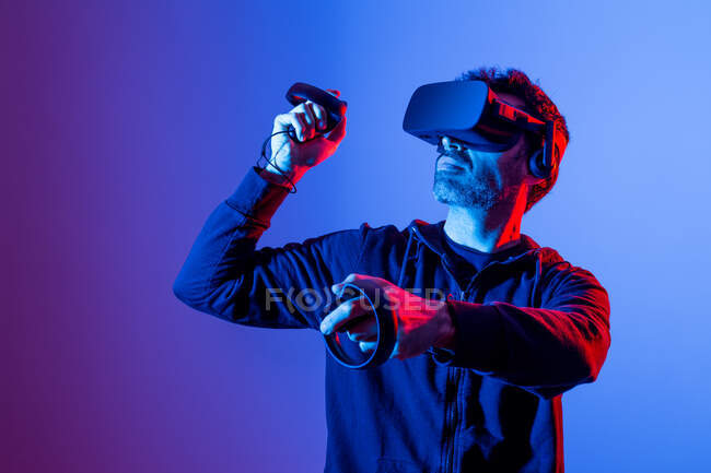 Anonymous unshaven male in hoodie and modern goggles with controllers and extended arm experiencing virtual reality — Stock Photo