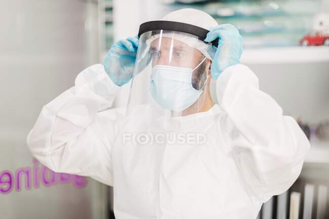 Positive male doctor with latex gloves and white uniform putting on face shield protective mask standing in modern medical office and looking at camera — Stock Photo