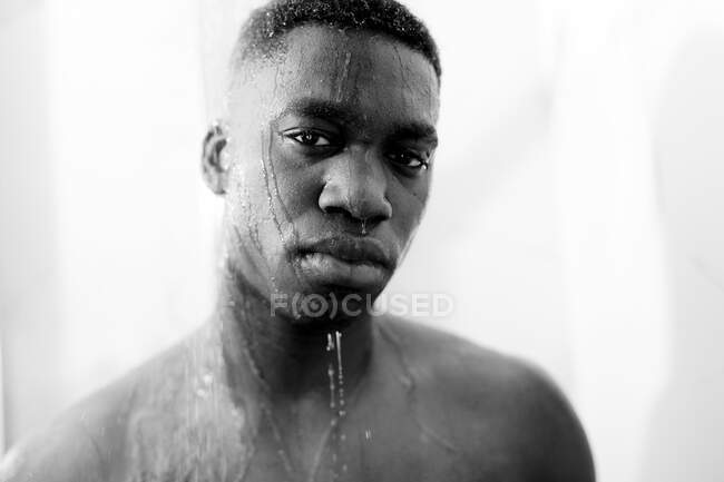 Noir et blanc de jeune homme noir sans émotion prenant une douche dans la salle de bain légère et regardant la caméra et l'eau sur le visage — Photo de stock
