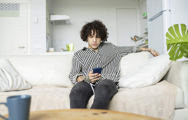 Молодой хипстер мужчина с кудрявыми волосами просматривает интернет на смартфоне во время отдыха на диване в комнате — стоковое фото