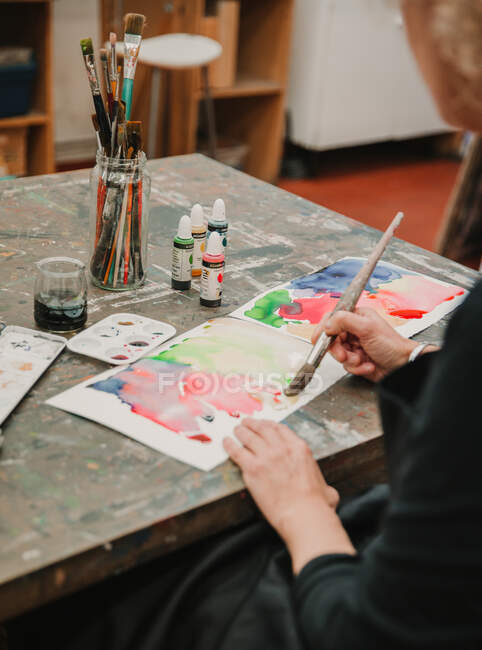 Corte a pintura artista feminina irreconhecível com aquarelas no papel enquanto estiver sentado à mesa no estúdio de arte — Fotografia de Stock