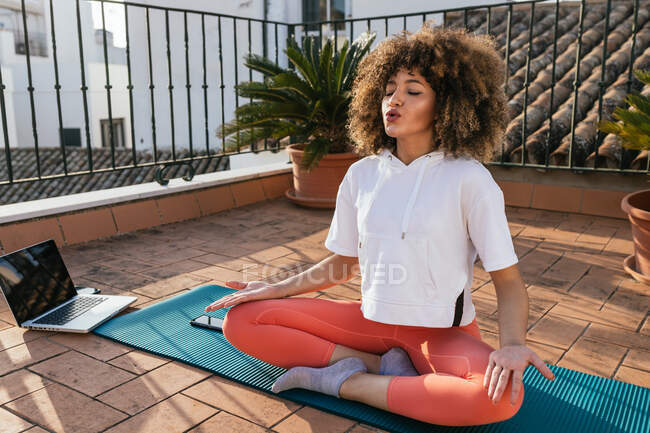 Tranquila mujer afroamericana sentada meditando con los ojos cerrados mientras practica yoga en la azotea - foto de stock