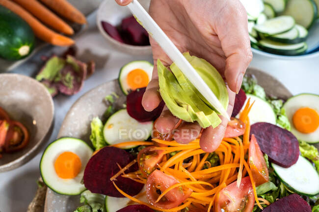 Desde arriba de la cosecha irreconocible persona cortar aguacate maduro mientras se prepara comida vegetariana con verduras variadas - foto de stock