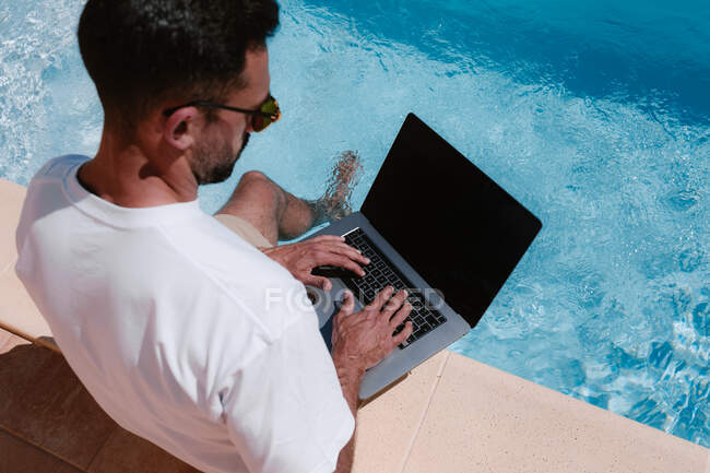 Dall'alto vista posteriore del freelance maschio in occhiali da sole seduto a bordo piscina e navigare netbook mentre si lavora a distanza sul progetto durante le vacanze estive — Foto stock